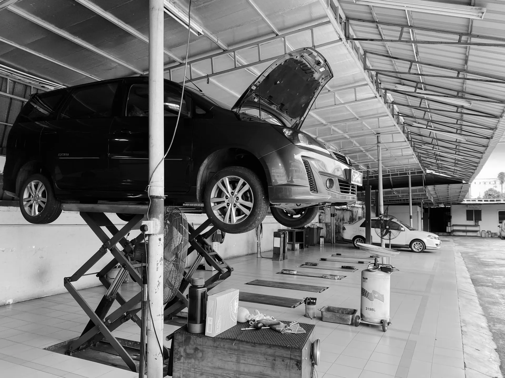 Black-and-white view of a family car on a scissor lift with its bonnet open in an open-sided workshop.