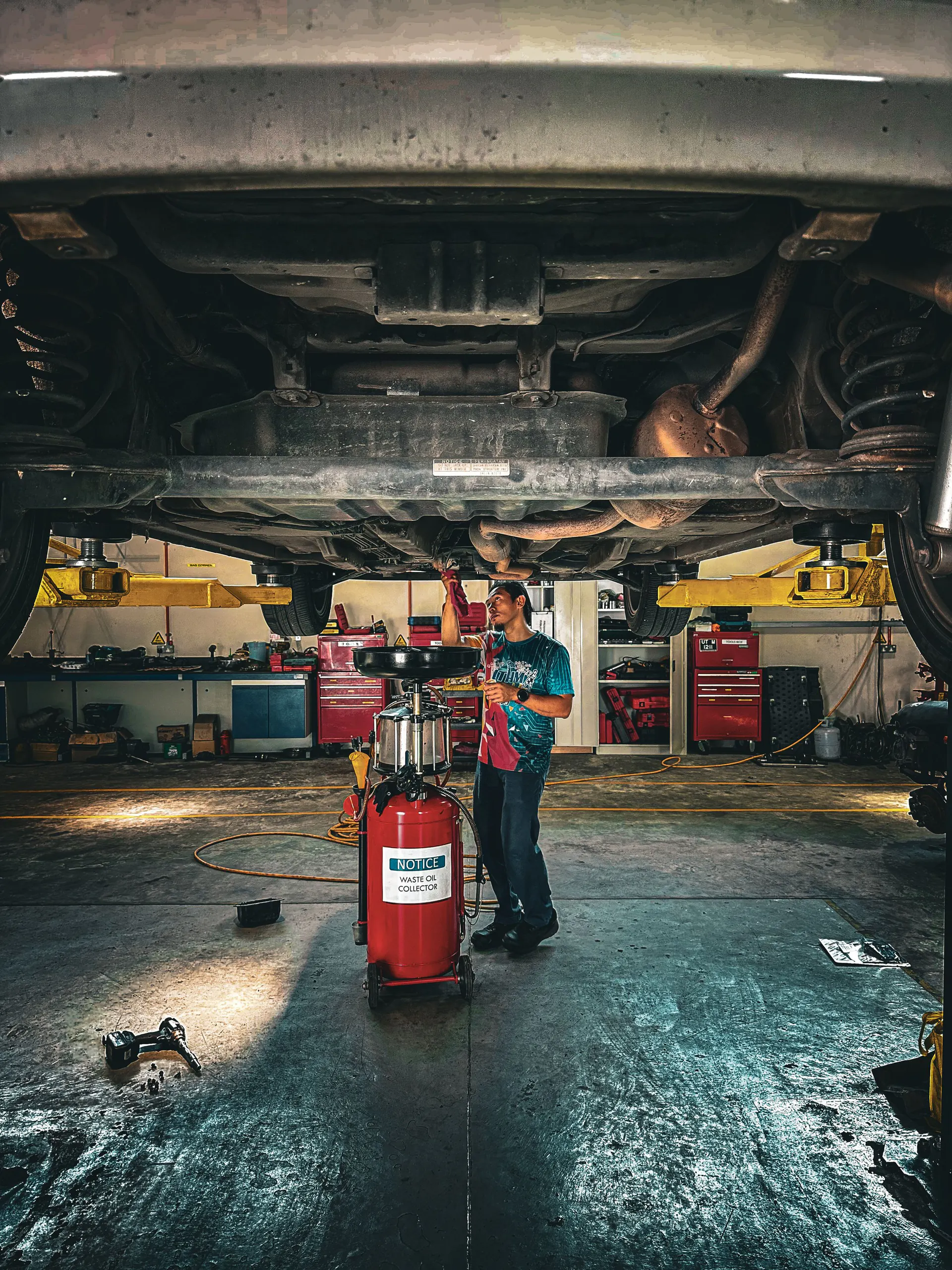 A mechanic works under a raised car beside a red waste-oil collector during an oil change.
