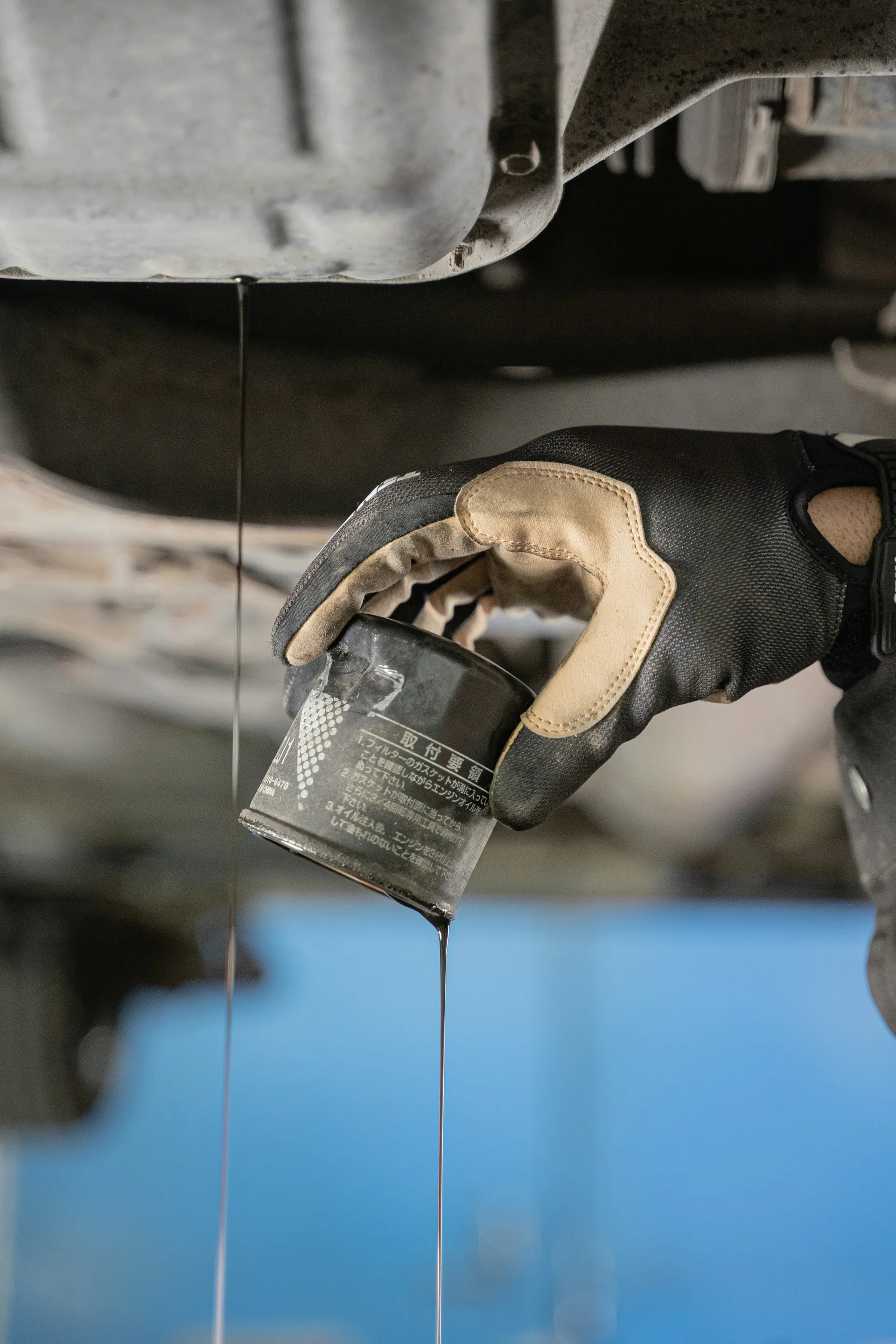 A gloved hand removes an old oil filter as used engine oil drips from under a car.
