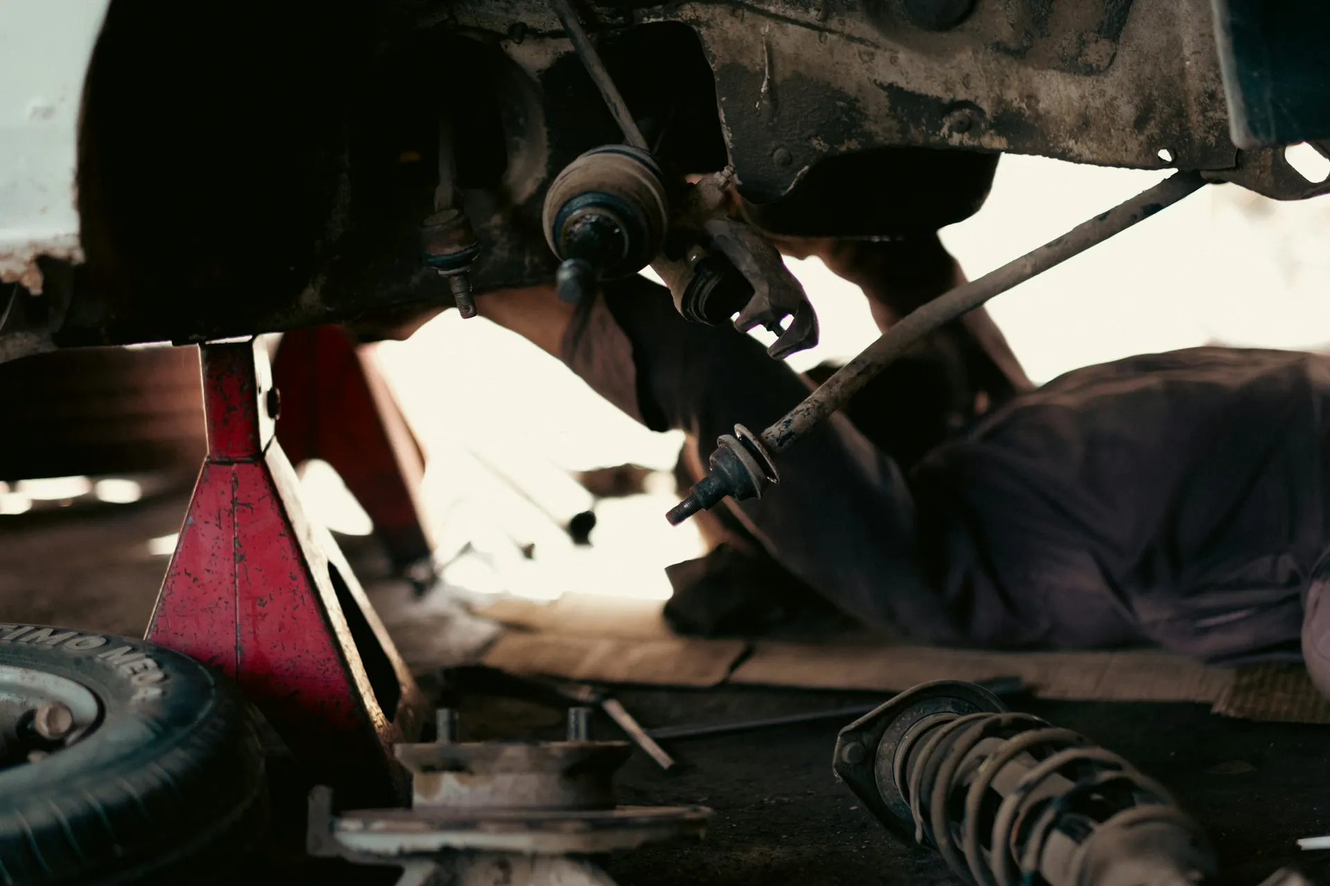 A mechanic lies under a car propped on a red jack stand, with suspension parts on the floor.
